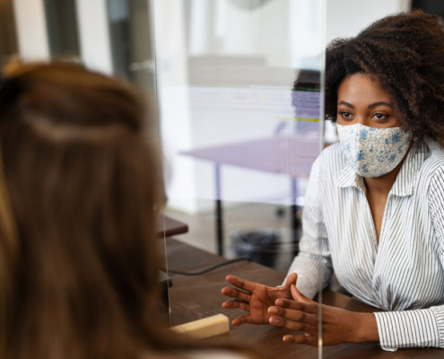 Female professional talking with colleague with a sneeze guard between them. Businesswomen wearing protective face masks for protection against virus while working in office.