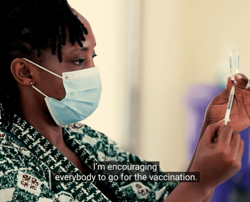 Screenshot from a World Health Organization video showing a healthcare worker in Ghana wearing a mask and preparing a COVID-19 vaccine. The caption reads: "I'm encouraging everybody to go for the vaccination."