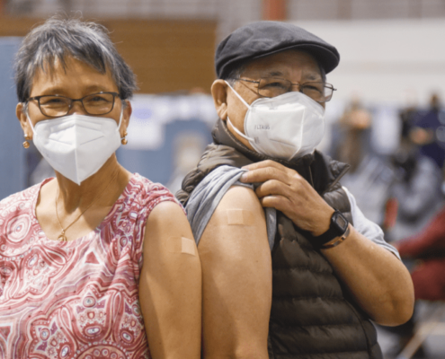 Two people wearing masks and showing where they received a COVID-19 vaccine.